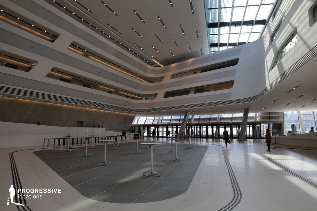 This expansive atrium showcases a striking contemporary design with flowing, organic lines and a multi-tiered structure. The curved balconies and recessed lighting create a sleek, futuristic aesthetic, while the use of white surfaces, glass panels, and metal accents enhances the space’s openness. A vast skylight floods the area with natural light, emphasizing the smooth contours and dynamic geometry of the ceiling and walls. The polished floors and minimalist furnishings contribute to the modern, high-tech atmosphere. This location is ideal for filming corporate settings, sci-fi environments, or high-end architectural visuals, suitable for genres like futuristic dramas, technology-focused narratives, or stylish commercial productions.