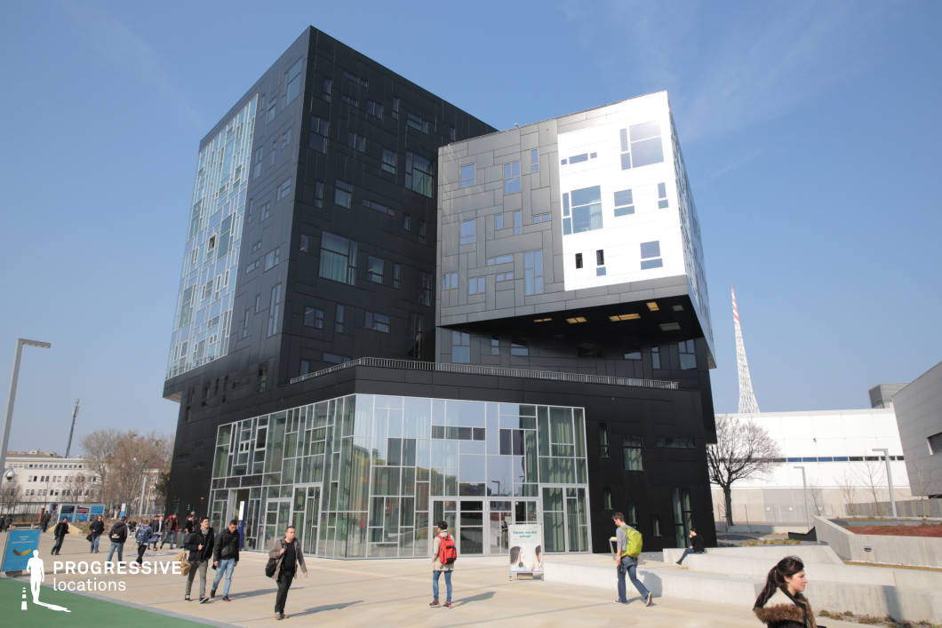 This striking contemporary building features a bold geometric design with a combination of black, glass, and metallic facades. The irregularly stacked structure creates a dynamic visual effect, with sections cantilevering over the lower floors. The extensive use of reflective and tinted glass panels enhances the futuristic aesthetic while allowing ample natural light inside. The surrounding area is open, with clean walkways and modern urban landscaping that integrates seamlessly with the structure’s sleek architecture. This location is ideal for filming corporate, sci-fi, or high-tech thriller scenes, as well as settings that require an avant-garde academic or research institution.