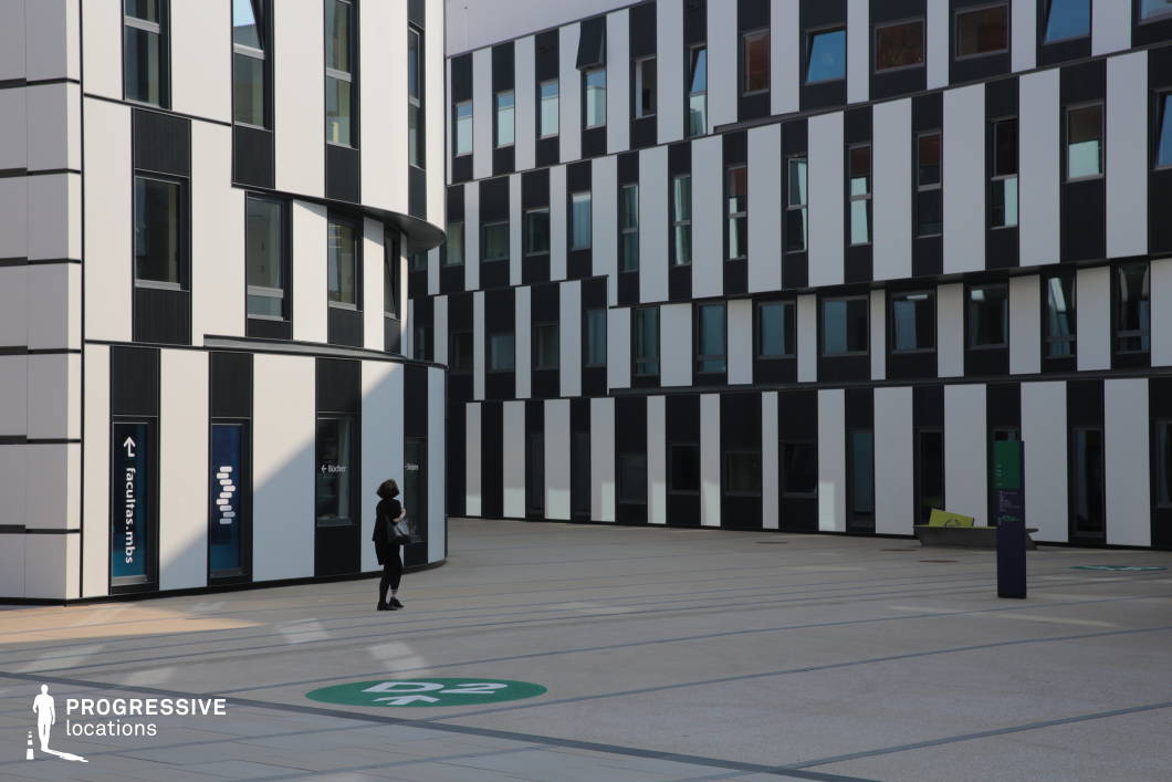 This modern architectural setting features a striking black-and-white striped facade with an irregular pattern, giving the buildings a bold, futuristic aesthetic. The slightly curved corner structure adds visual contrast to the otherwise angular design, while the numerous rectangular windows are arranged asymmetrically, contributing to a dynamic and unconventional look. The open courtyard is paved with neutral-colored tiles, marked with subtle geometric patterns and directional signage, enhancing its structured and organized atmosphere. The interplay of light and shadow on the facade emphasizes the depth and texture of the design. This location is ideal for filming corporate, sci-fi, or intellectual drama settings, offering a sleek, avant-garde backdrop suitable for narratives involving innovation, academia, or high-tech urban environments.