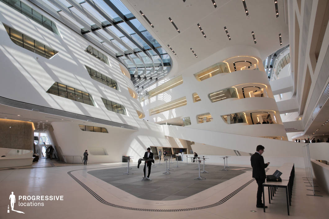 This striking atrium showcases an avant-garde architectural design, featuring sweeping curves, asymmetrical balconies, and expansive skylights that flood the space with natural light. The white, fluid-like walls contrast with sleek glass windows and warm recessed lighting, creating a futuristic and high-tech aesthetic. The open-concept layout, with multiple levels interconnected by curved walkways, adds depth and movement to the scene. Minimalist furniture and clean geometric flooring enhance the ultramodern ambiance. This location is ideal for science-fiction films, corporate thrillers, or high-concept dramas, serving as a backdrop for innovative technology firms, elite institutions, or utopian/dystopian worlds.