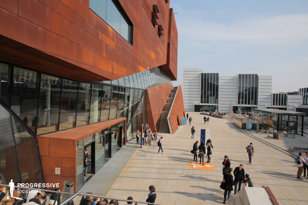 This striking urban setting features a bold architectural design, dominated by a rust-colored angular structure with large glass facades that reflect the surrounding environment. The dynamic geometry of the building contrasts with the sleek, modern white and black buildings in the background, creating a visually engaging space. The open plaza, paved with light-colored tiles, provides a spacious and inviting environment, enhanced by integrated staircases and elevated walkways. The mix of industrial and minimalist design elements makes this location ideal for films centered on academia, futuristic narratives, or corporate and innovation-driven stories, offering a sleek and progressive backdrop.