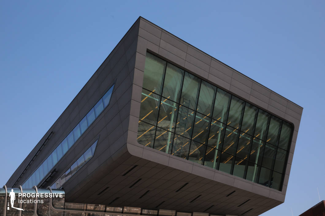 This striking modern building features a bold cantilevered design, extending outward with a sharp angular form. The exterior is clad in dark grey panels, contrasting with the large floor-to-ceiling glass windows that offer a glimpse into the illuminated interior. The geometric precision of the structure is emphasized by clean lines and sharp edges, creating a futuristic aesthetic. The building's elevated mass appears to defy gravity, giving it a dynamic and avant-garde presence. This location is ideal for filming scenes that require a high-tech, corporate, or dystopian setting, making it suitable for science fiction, thrillers, and architectural documentaries. The dramatic overhang and reflective glass surfaces provide excellent opportunities for visually compelling compositions and striking cinematography.