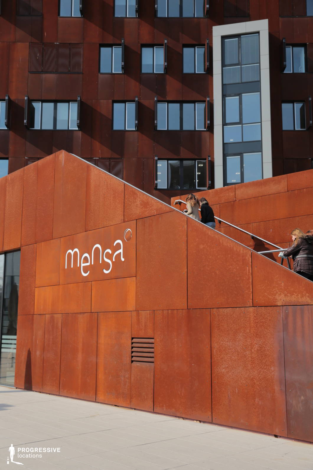 This contemporary building features a striking rust-colored corten steel facade, characterized by its weathered texture and industrial aesthetic. The angular stairway, integrated seamlessly into the structure, adds a dynamic geometric element, guiding the eye toward the upper levels. The facade is punctuated by large, asymmetrically placed windows with black metal frames, creating a contrast between the reflective glass and the raw steel. The interplay of light and shadow on the building’s surface enhances its depth and visual interest. This location is ideal for filming urban, architectural, or futuristic settings, offering a distinctive industrial-chic atmosphere suitable for corporate, academic, or dystopian-themed productions.