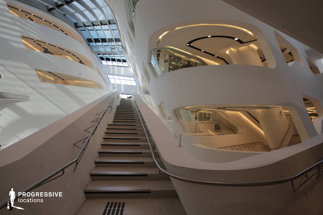 This striking modern interior features an open, multi-level layout characterized by smooth, curvilinear white walls and expansive glass panels that allow natural light to flood the space. A long, ascending staircase with sleek metal railings leads the eye upward, emphasizing the sense of scale and openness. The design incorporates asymmetrical cutouts and curved balconies with integrated lighting, creating a futuristic and dynamic aesthetic. The interplay of light and shadow enhances the architectural depth, making this location ideal for filming sci-fi, high-tech corporate settings, or avant-garde conceptual sequences, offering a sleek and visually immersive backdrop.