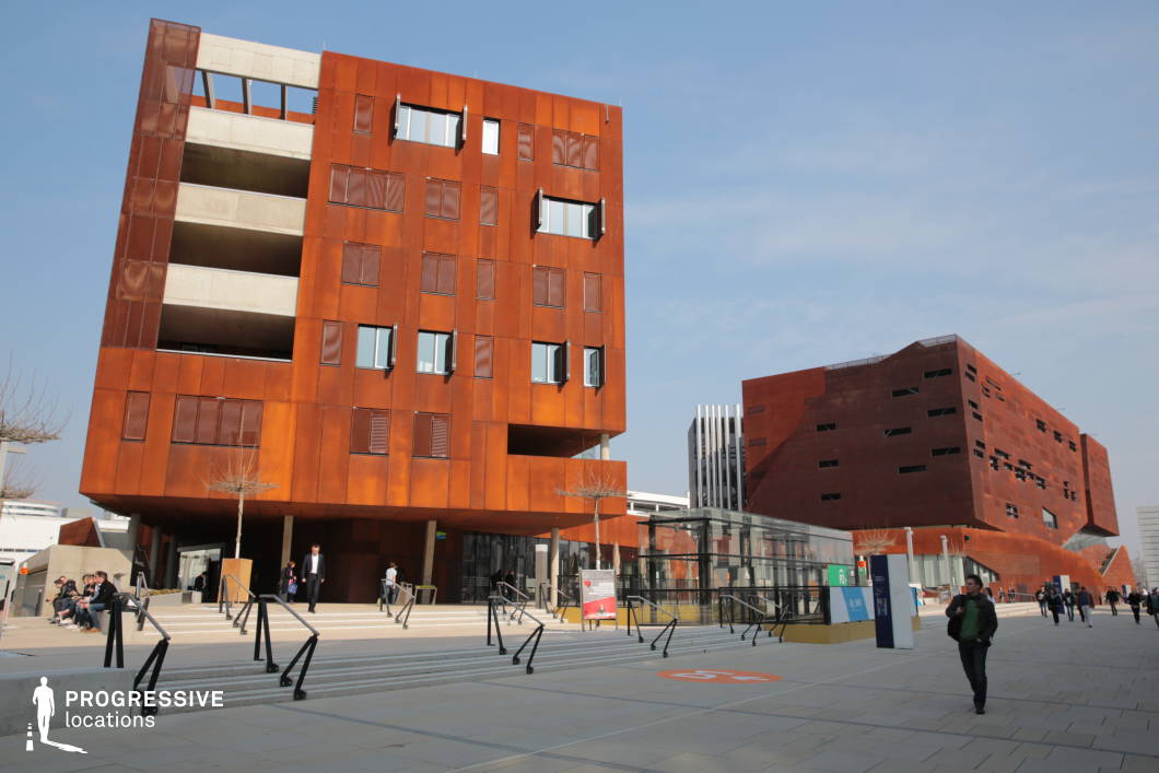 This architectural complex features striking corten steel-clad buildings with a rust-like patina, creating an industrial yet contemporary aesthetic. The angular and asymmetrical structures, with irregularly placed windows and deep recesses, give a dynamic, sculptural quality to the space. The open plaza in the foreground is paved with large, neutral-toned tiles and features multiple levels connected by staircases and ramps, enhancing accessibility and spatial layering. Glass elements, including a modern walkway enclosure, introduce transparency and contrast with the dominant warm-toned metal facades. This location is ideal for futuristic, dystopian, or corporate-themed film settings, providing a visually rich environment suitable for intellectual, institutional, or high-tech corporate narratives.