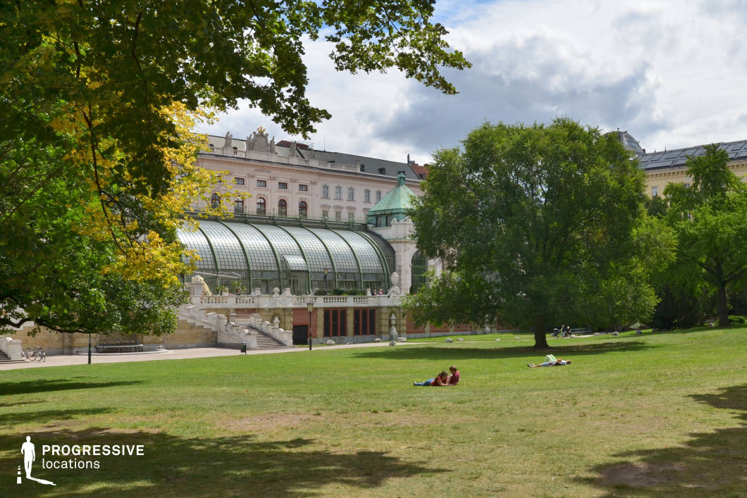 This picturesque scene features a grand glasshouse with a curved metal and glass structure, seamlessly integrated into a historic building adorned with classical architectural details. The elegant staircases, ornate balustrades, and arched windows add a refined European charm to the setting. The foreground consists of an expansive, well-maintained lawn bordered by mature trees, creating a tranquil and inviting atmosphere. The combination of historic architecture and lush greenery offers a versatile backdrop suitable for period films, romantic dramas, or intellectual and contemplative scenes, providing a perfect balance of sophistication and natural beauty.