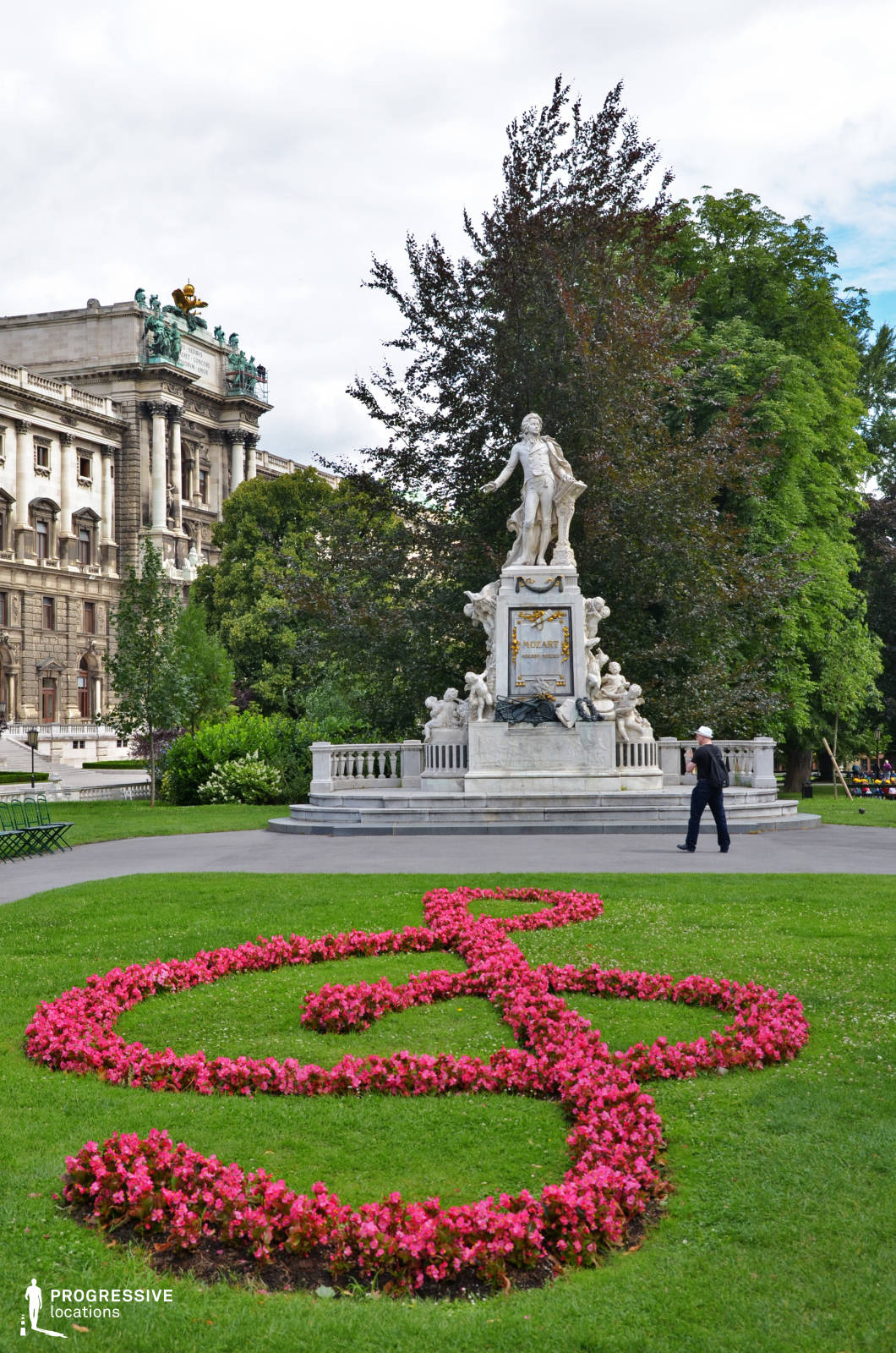 The image showcases a grand neoclassical monument dedicated to Mozart, set within a lush, manicured park. The white marble statue stands on an ornate pedestal adorned with golden details and intricate sculptural elements, exuding historical and artistic significance. In the foreground, a vibrant floral arrangement in the shape of a treble clef adds a striking visual contrast against the deep green grass. Towering trees frame the scene, providing depth and a natural backdrop. In the background, an opulent historic building with grand columns and sculptural decorations reinforces the location's classical elegance. This setting is ideal for historical and period dramas, as well as refined artistic productions. It provides a sophisticated atmosphere for scenes involving classical music, cultural heritage, or intellectual discourse.