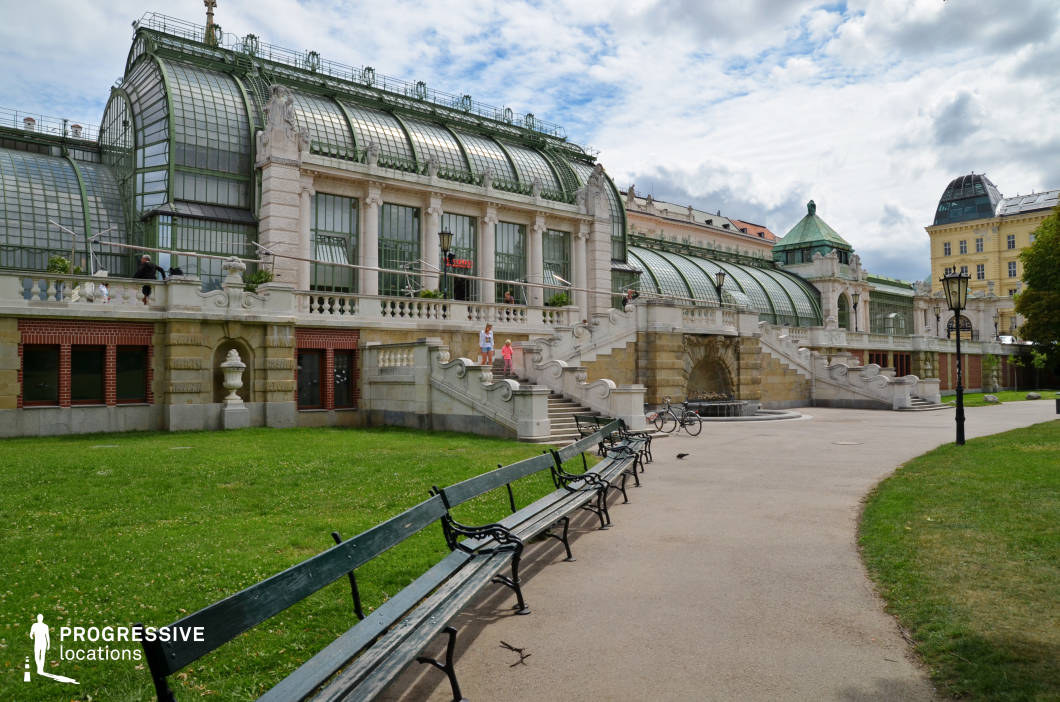 This visually striking location features a historic greenhouse with an ornate, neoclassical façade and large glass panels framed by intricate metalwork. The grand stone staircase with sculpted balustrades adds a regal touch, leading to an elevated terrace that enhances the sense of scale. The surrounding park includes well-maintained green lawns, paved walkways, and classic wrought-iron benches that provide a picturesque foreground. The architectural contrast between the greenhouse’s delicate glass structure and the solid stone elements creates a refined yet inviting atmosphere. This setting is ideal for filming period dramas, romantic scenes, or sophisticated urban sequences, offering a blend of historic grandeur and lush greenery.