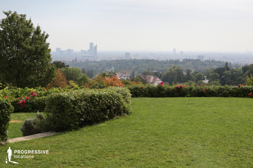 This elevated garden setting provides a sweeping panoramic view of a cityscape in the distance, blending urban architecture with lush greenery. The foreground features a well-manicured lawn bordered by neatly trimmed hedges and vibrant floral elements, creating a structured yet natural aesthetic. The gradual transition from the garden’s cultivated landscape to the dense treetops and scattered rooftops below adds depth, while the distant skyline with modern high-rises establishes a striking contrast between nature and urbanization. This location is ideal for reflective or dramatic scenes, romantic moments, and establishing shots in films spanning genres such as drama, romance, or introspective storytelling.
