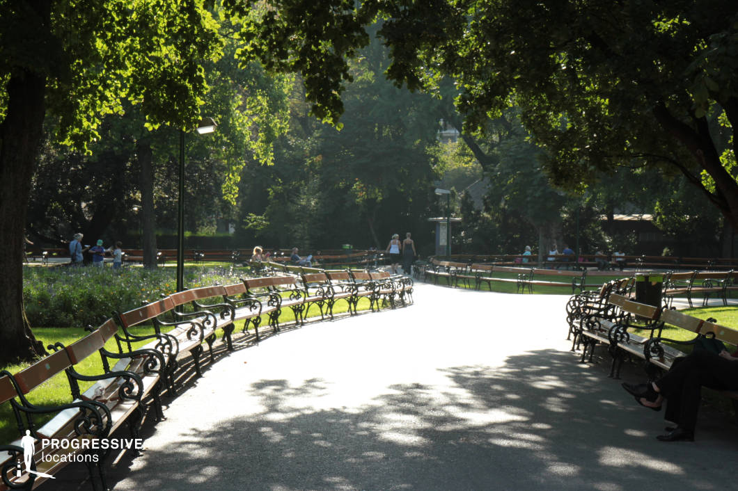 This serene park setting features a gracefully curved pathway lined with classic wooden benches with intricate wrought iron detailing. Towering trees with lush green foliage frame the scene, casting dappled shadows on the well-maintained pavement. The surrounding landscape is filled with dense greenery and flower beds, creating a tranquil urban retreat. Traditional lampposts add an elegant touch, complementing the park’s historic ambiance. The depth of the perspective and the natural enclosure formed by the trees make this location ideal for filming contemplative scenes, romantic encounters, or introspective moments in dramas, period films, or artistic cinematography focused on urban nature.