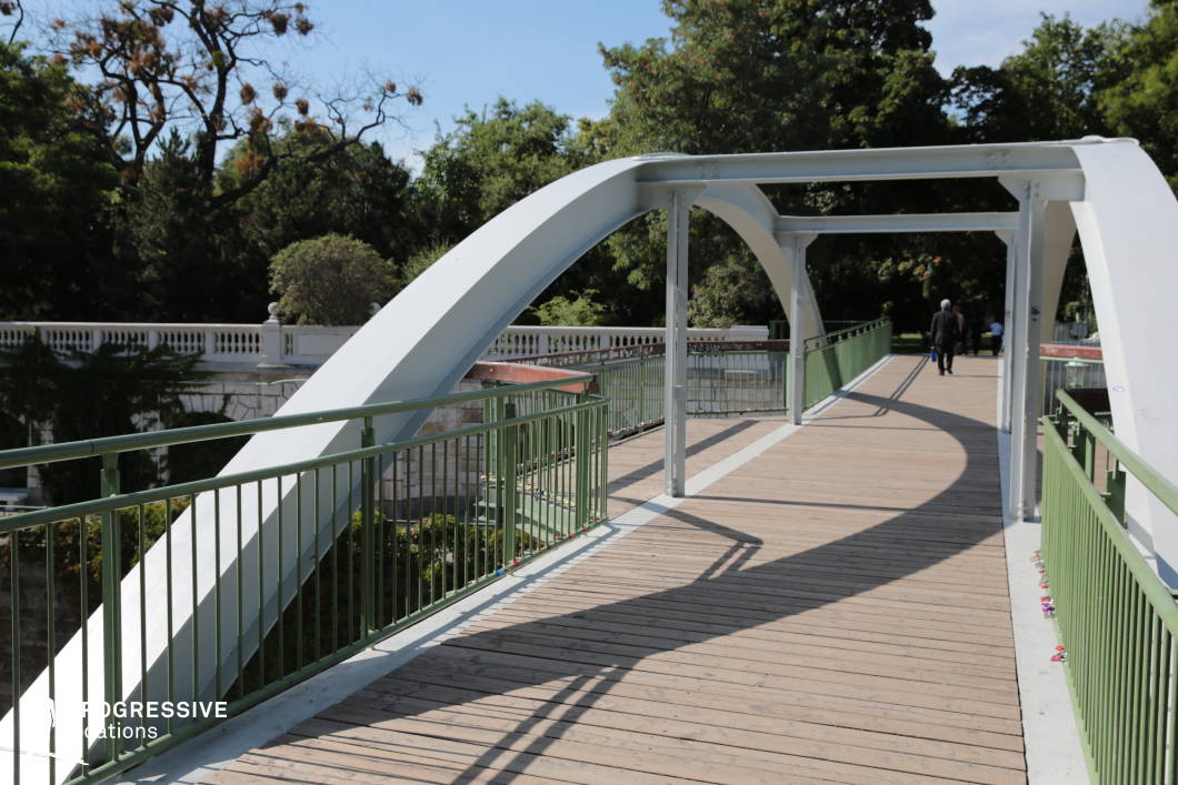 This location showcases a sleek pedestrian bridge with a white metal arch structure and green railings, set against a lush park landscape. The wooden walkway adds a natural texture, contrasting with the industrial feel of the steel framework. The bridge spans over a water feature, with a decorative white balustrade visible in the background, adding a classical touch to the otherwise modern design. The combination of geometric lines, shadows, and open space makes this location ideal for dynamic tracking shots, suspenseful thriller scenes, or architectural and urban-themed film projects. Its mix of natural and contemporary elements also suits romantic or contemplative sequences.