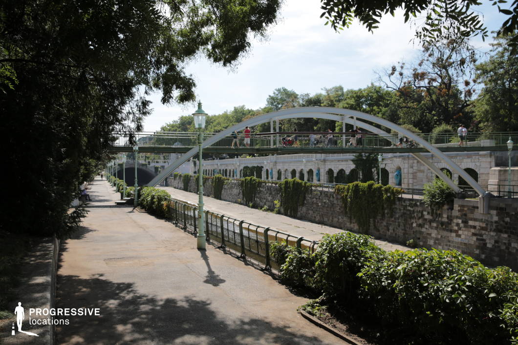 This scenic urban setting features a gracefully arched pedestrian bridge spanning a narrow canal, surrounded by lush greenery and historic stonework. The canal, lined with ivy-covered stone walls and ornate lampposts, creates a balanced blend of natural and architectural elements. The gently curving walkway along the waterway offers depth and perspective, framed by overhanging trees that add a serene atmosphere. The bridge’s sleek metal structure contrasts with the classical details of the stone arches beneath it, providing a visually dynamic composition. This location is ideal for romantic films, reflective dramas, or atmospheric cityscape sequences, offering a picturesque yet timeless urban backdrop.