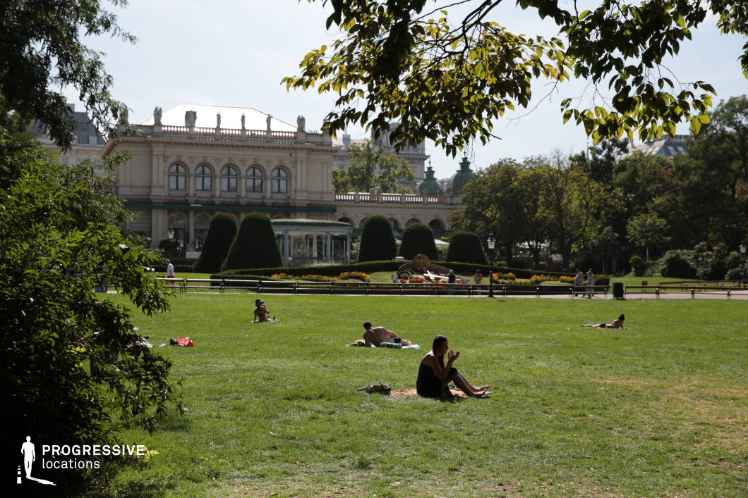 This scenic city park blends lush greenery with refined architecture, featuring an expansive lawn framed by manicured hedges and vibrant flower beds. In the background, a grand neoclassical building with arched windows, decorative balustrades, and statues atop its roof adds a sense of sophistication and old-world charm. The symmetrical landscaping, including carefully shaped topiaries and a central decorative element, enhances the orderly yet inviting atmosphere. The park’s open space, lined with classic benches and tree-shaded walkways, creates a versatile setting suitable for filming leisurely urban scenes, romantic encounters, or elegant period pieces. Its combination of nature and historic architecture makes it ideal for productions requiring a refined, European-style backdrop.