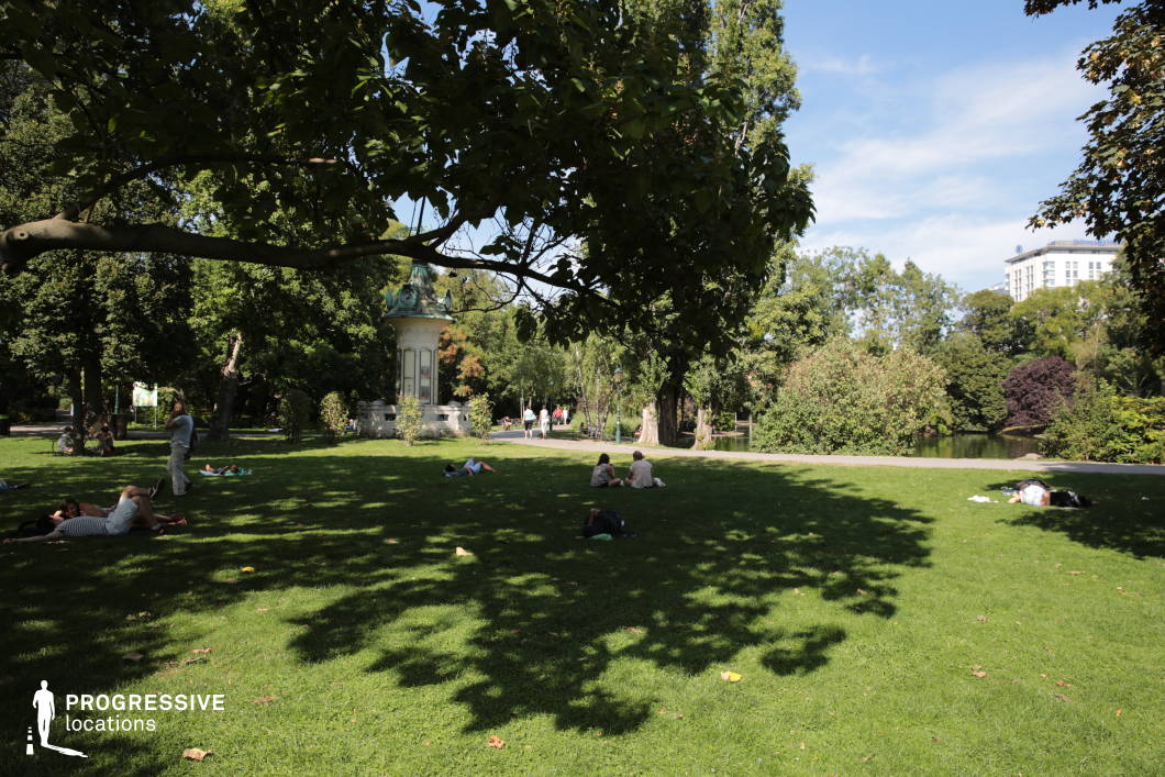 This serene urban park features an expansive, well-maintained lawn framed by mature trees that cast intricate shadows, adding depth and contrast to the setting. In the background, a reflective lake is bordered by a diverse array of trees and shrubs, creating a layered natural composition that enhances the sense of tranquility. The combination of open green space and the water's edge provides a visually balanced backdrop, making this location ideal for filming peaceful outdoor scenes, contemplative moments, romantic walks, or casual city life sequences that require a natural yet structured environment.