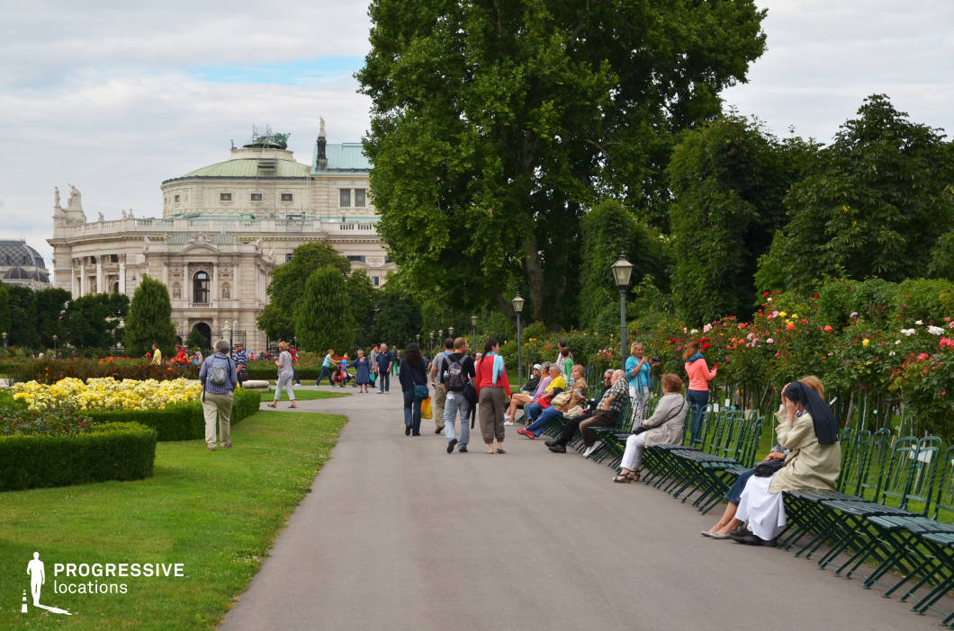 This location features a meticulously maintained park with vibrant flower beds, neatly trimmed hedges, and a paved walkway lined with classic green metal benches. Towering trees provide shade, while vintage-style street lamps contribute to the park's old-world charm. In the background, an ornate neoclassical building with intricate detailing, statues, and a green copper roof adds grandeur to the scene. The combination of lush greenery and stately architecture creates an elegant and serene atmosphere, making this an excellent setting for filming period dramas, historical narratives, or romantic scenes. The park’s structured layout and classical aesthetic also suit documentary projects and high-end fashion shoots.