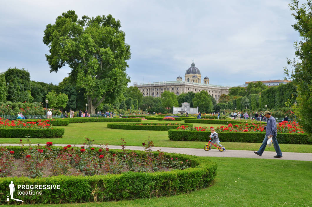 This meticulously landscaped garden features geometrically arranged flower beds filled with vibrant red roses, neatly trimmed hedges, and expansive green lawns. A grand historic building with a domed roof and ornate architectural details dominates the background, adding a sense of grandeur and cultural significance to the scene. Towering trees frame the park, creating a natural canopy that enhances the sense of depth and tranquility. Paved walkways weave through the lush greenery, offering a structured yet inviting atmosphere. This location is ideal for filming period dramas, romantic sequences, or elegant city strolls, providing a refined and picturesque setting that blends nature with historic architecture.