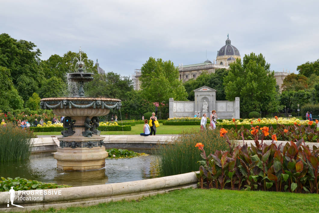 This lush, meticulously landscaped garden features an ornate, multi-tiered stone fountain as its focal point, adorned with sculpted figures and intricate detailing. Surrounding the fountain, a circular water basin adds a tranquil, reflective quality to the scene. The garden is structured with neatly arranged flower beds showcasing vibrant red and yellow blooms, alongside well-manicured hedges that define the pathways and green spaces. In the background, a grand neoclassical monument with a sculpted relief stands prominently, framed by lush trees that create depth and a sense of enclosure. Beyond the greenery, a stately domed building with intricate architectural details enhances the historical ambiance. This location is ideal for period dramas, romantic scenes, and elegant commercial shoots, offering a refined, European aesthetic with a timeless atmosphere.
