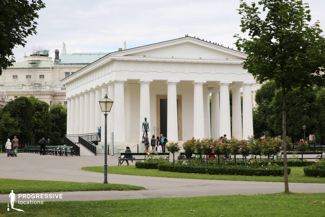 This striking neoclassical temple stands in the center of a manicured park, surrounded by lush greenery and symmetrically arranged flower beds. Its pristine white facade, adorned with evenly spaced Doric columns, creates a strong sense of balance and grandeur. The triangular pediment and smooth architectural lines contrast elegantly with the historical buildings in the background, adding depth to the scene. Paved walkways and traditional lampposts enhance the classical ambiance, making this location ideal for period films, philosophical dramas, or historical documentaries. Its serene yet monumental presence provides a timeless setting for intellectual dialogues, political intrigue, or reflective character moments.