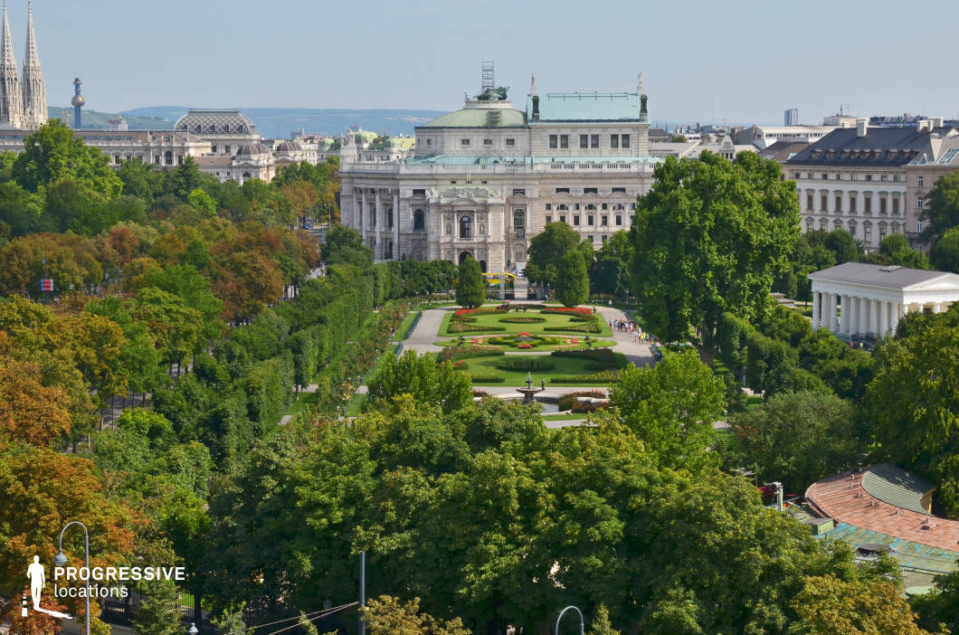 This image showcases an elegant, symmetrically designed garden surrounded by lush greenery, leading to an opulent neoclassical building with intricate detailing, statues, and a green copper roof. The foreground is dominated by dense tree canopies, framing the structured pathways and manicured flower beds that extend toward the central architectural focal point. To the right, a white, columned classical structure adds a contrasting element to the scene. The background features a mix of historic and modern architecture, with spires and domed rooftops visible beyond the treetops. The location's grandeur, symmetry, and balanced composition make it ideal for filming period dramas, historical epics, or romantic sequences, offering a refined and cinematic atmosphere.