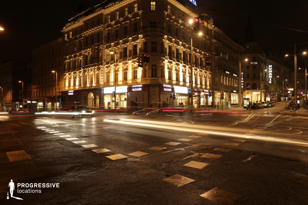 This urban street scene at night features a bustling intersection with traffic moving through a well-lit area. The architecture is dominated by a grand corner building with classical detailing and modern storefronts illuminated by bright lights, contributing to a vibrant city atmosphere. The traffic lights are visible, with streaks of light from moving vehicles adding dynamic energy to the scene. The surrounding buildings reflect a mix of historic and contemporary design elements, with neon signs adding a modern touch. The road surface appears wet, enhancing the reflective qualities of the lights. This location, with its dramatic blend of urban architecture, lighting, and street activity, is ideal for shooting urban thrillers, action scenes, or city life portrayals. Its visual appeal makes it versatile for various genres like crime dramas, romantic city scenes, or commercial advertisements.