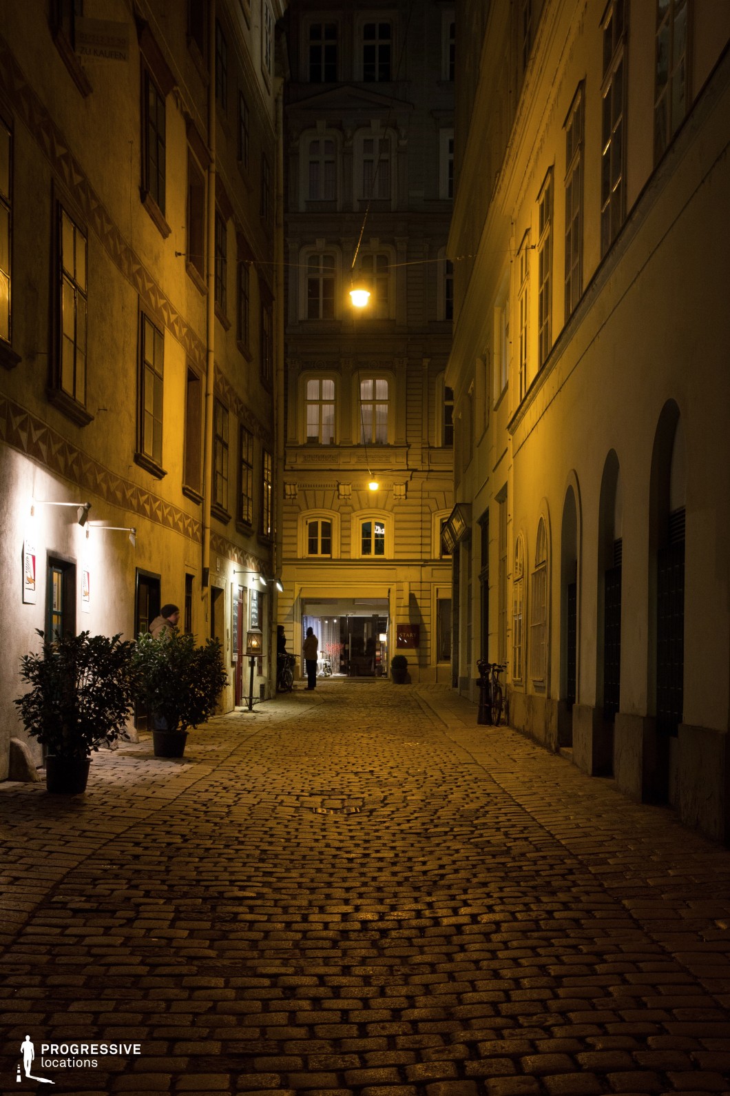This narrow cobblestone alley, illuminated by warm street lamps, showcases the charm of a historic city center. The buildings on either side feature classical architecture, with detailed facades and arched windows. The path, made of uneven cobblestones, adds texture and depth to the scene, creating a feeling of intimacy and solitude. The lighting casts a soft, yellow glow, enhancing the nostalgic atmosphere of the location. The alley is tucked away, offering a sense of privacy while remaining part of the lively city environment. This location is ideal for intimate, suspenseful, or romantic scenes, and is well-suited for genres like drama, thriller, or mystery. It offers a perfect backdrop for shooting night scenes with its atmospheric lighting and historic urban feel.