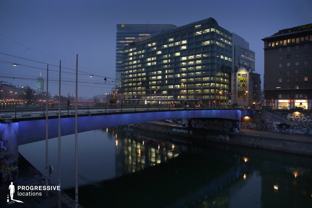 This image captures a modern urban landscape along a canal, with a sleek, contemporary office building dominating the background. The illuminated windows of the building reflect on the calm waters of the canal, adding a dynamic visual contrast to the structure. A bridge, lit with soft blue lighting, crosses the water, providing a focal point for the composition. Graffiti-covered walls along the canal’s edge hint at the area’s urban character. This location, with its mix of modern architecture, canal, and street art, is ideal for filming genres such as urban dramas, thrillers, or modern action sequences. The setting offers both a vibrant city backdrop and opportunities for dramatic shots of the waterway and architecture.
