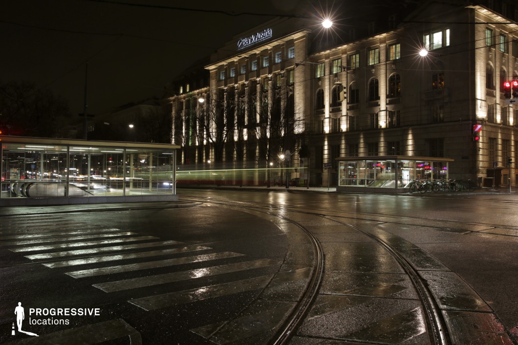 This nighttime scene features an urban intersection, with a sleek modern metro station shelter standing at the forefront. The surrounding environment is illuminated by streetlights that highlight the area, creating a reflective, almost cinematic effect. In the background, the grand facade of a historical building is bathed in warm light, contrasting with the modern infrastructure in the foreground. The tram tracks curving through the street add dynamic lines to the composition. This location, with its mix of contemporary and classical architecture, is ideal for filming urban dramas, thrillers, or action scenes. The intersection offers a great opportunity for motion shots involving trams or people, and the lighting enhances the visual impact of the scene.