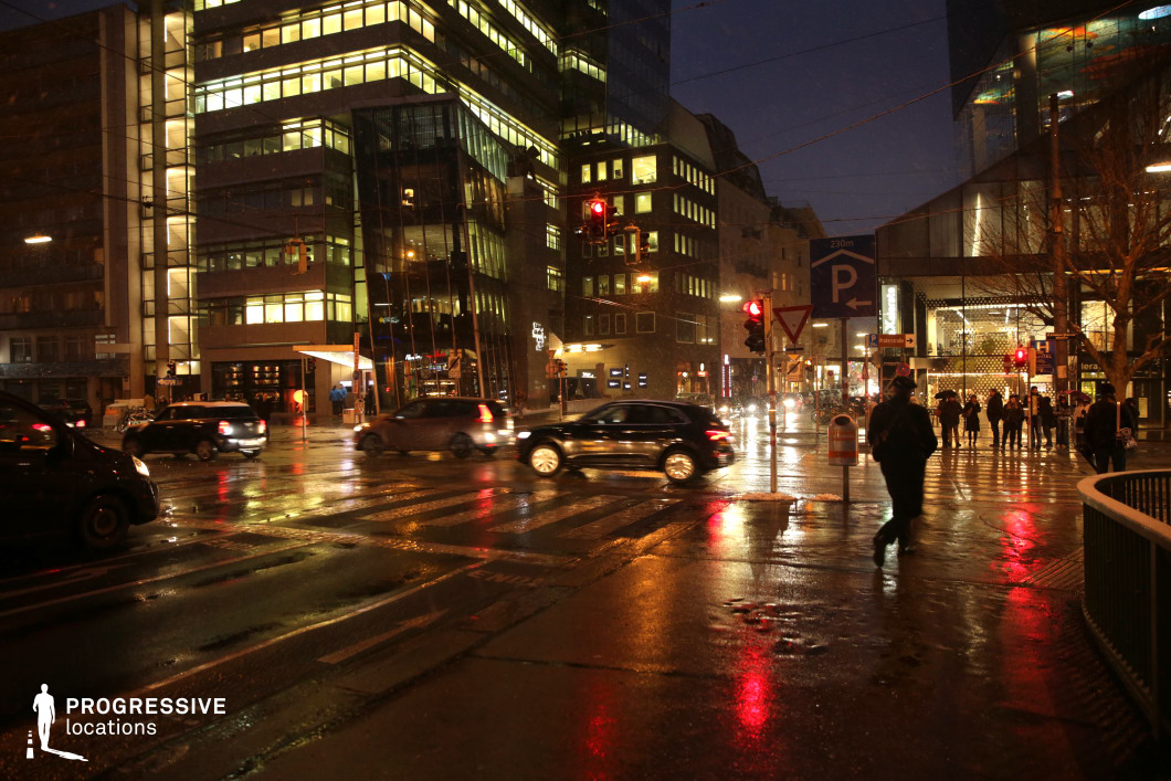 This nighttime scene showcases a busy urban intersection with modern glass buildings that dominate the skyline. The sleek architecture of the surrounding buildings is beautifully illuminated, creating a striking contrast with the dark sky. The reflective surfaces of the wet street add a cinematic touch, enhancing the glow of neon signs and streetlights. The crosswalk lines are visible, leading into the heart of the scene, where the urban design and lighting play a prominent role. This location is perfect for films requiring a modern, dynamic city backdrop, ideal for corporate, thriller, or action scenes that highlight sleek, contemporary environments.
