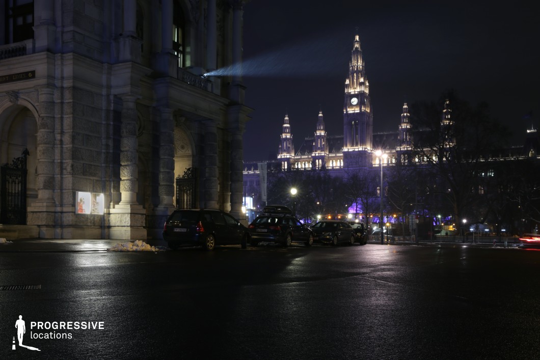 The image showcases a grand urban scene at night, where majestic architecture stands illuminated against a deep blue sky. In the foreground, a large building with stone pillars and classical detailing is paired with modern elements, such as light projection equipment. The central focus is the grand clock tower of a distant government building, outlined by dramatic lighting, creating a sharp contrast with the dark surroundings. The glossy street surface reflects the lights, enhancing the atmosphere. The scene combines historical and contemporary architecture, highlighted by stark contrasts in lighting and shadows. This location offers a dynamic backdrop for a variety of filming genres, including historical dramas, thrillers, or urban dramas, with its cinematic lighting and impressive architectural elements. The large open spaces and iconic city landmarks make it ideal for wide shots and establishing scenes.