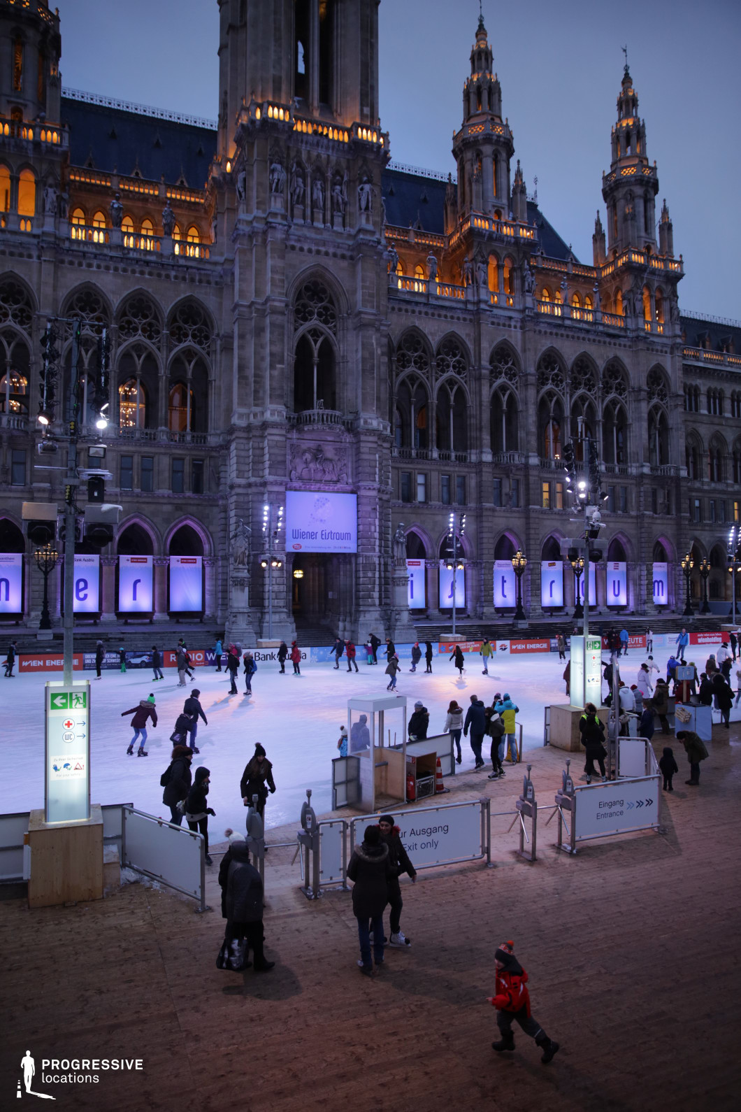 This vibrant winter scene takes place at Rathausplatz, featuring a large outdoor ice rink in front of the majestic, Gothic-style Rathaus (City Hall) building. The illuminated structure's intricate architecture stands out with warm lights highlighting its spires and arches. The ice rink is filled with skaters, creating a lively atmosphere, while festive lighting adds a touch of magic to the scene. The cobblestone ground around the rink and the surrounding buildings contribute to the historical ambiance. This location is perfect for showcasing winter scenes in films, particularly in romantic, family-oriented, or holiday genres. Its festive and grand setting is ideal for capturing winter activities, social gatherings, or holiday celebrations.
