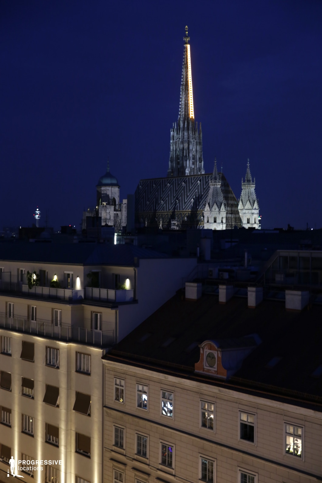 This nighttime scene offers a stunning view of the St. Stephen's Cathedral spire, illuminated and standing tall above the rooftops of the surrounding buildings. The cathedral's intricate gothic architecture is highlighted by the lights, with its iconic spire piercing the dark sky. Below, the surrounding rooftops are bathed in soft lighting from street lamps and window lights, creating a warm, urban atmosphere. The contrast between the sharp, detailed spire and the more uniform city rooftops emphasizes the grand scale of the cathedral. This location is perfect for cinematic city shots, ideal for period dramas, historical narratives, or establishing shots in romantic or architectural films, offering a beautiful, timeless backdrop of the city.
