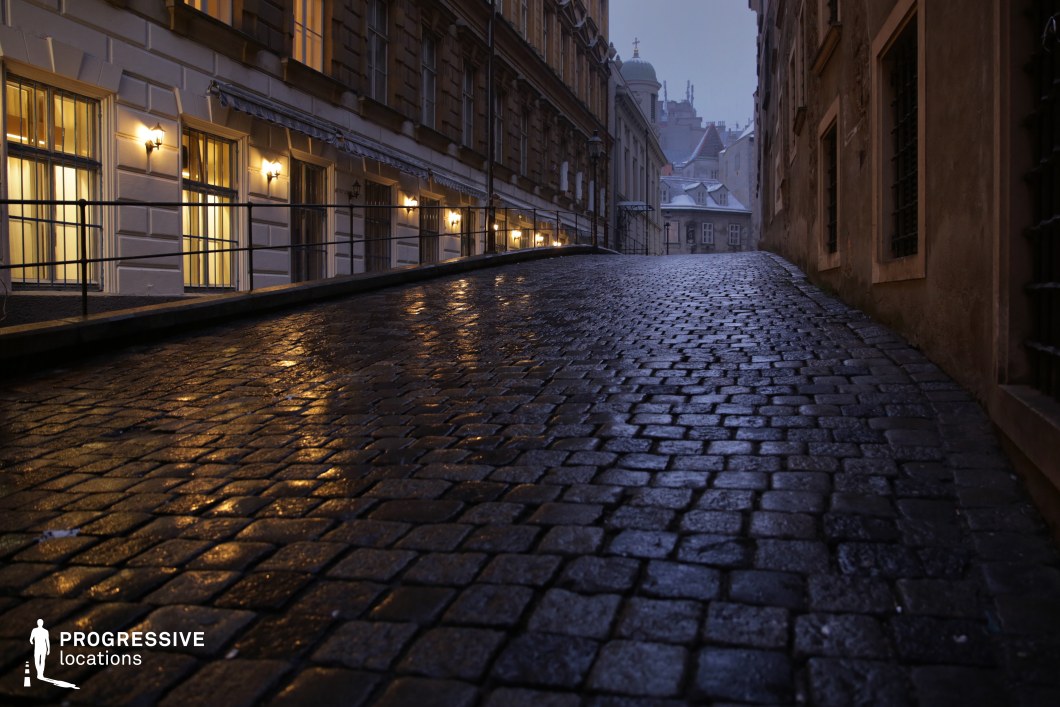 This narrow, uphill cobblestone street exudes an old-world charm, with the wet stones reflecting the warm glow of streetlights. The architecture on either side consists of historic buildings, with large windows and architectural details such as wrought-iron railings. The scene is illuminated by soft yellow lighting, casting a calm yet mysterious atmosphere. In the distance, the fading light of dusk adds a sense of anticipation to the scene. This location, with its historic ambiance and dramatic lighting, is ideal for films that require a moody, suspenseful, or eerie atmosphere. It is perfect for genres like mystery, thriller, or horror, offering an ideal setting for dark alley or secretive confrontations.