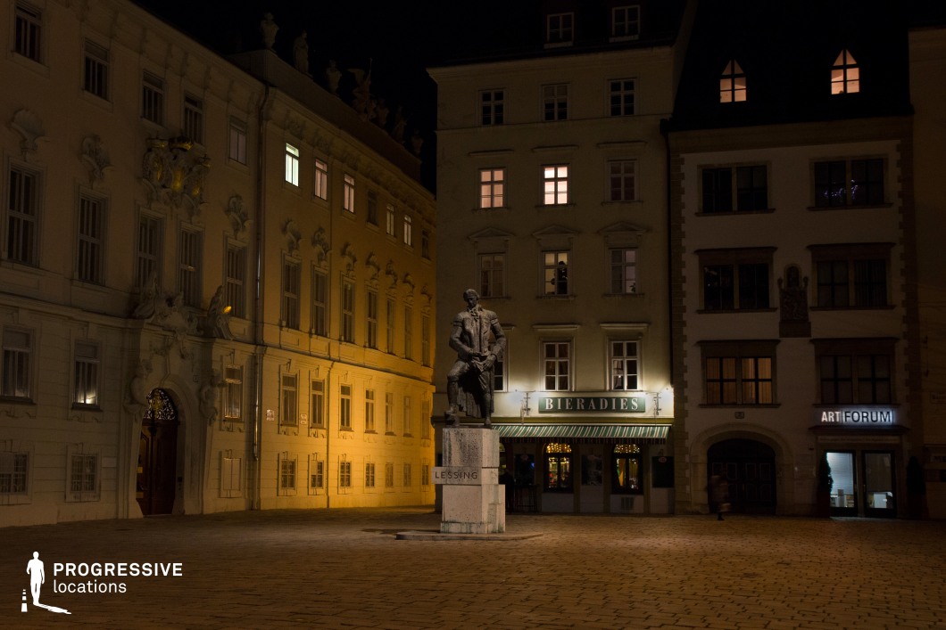 This image features a quiet cobblestone square illuminated by warm streetlights, with a striking statue of a historical figure at its center. The surrounding buildings, showcasing elegant baroque architecture, add to the square's charm, with their detailed facades and large windows. The contrast between the dark night sky and the soft golden lighting on the buildings creates a sophisticated and timeless atmosphere. This location, with its historical significance, elegant architecture, and cinematic lighting, is ideal for filming period dramas, historical films, or scenes requiring an elegant, classical backdrop. The open square and surrounding buildings offer opportunities for both wide establishing shots and close-ups of architectural details.