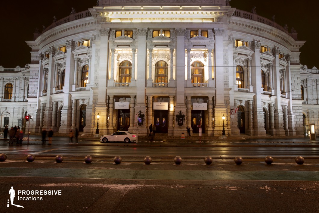 The photograph showcases a grand classical theatre building, illuminated by soft, warm lighting that highlights its detailed stone façade and intricate architectural elements. The symmetrical design of the building, with grand arches and ornate windows, creates a sense of elegance and historical grandeur. The surrounding street is quiet, with reflections on the wet pavement from the street lamps and the theatre's lighting. The contrast between the bright, well-lit structure and the shadowed surroundings enhances the building’s imposing presence. This location, with its refined architectural features and atmospheric lighting, is perfect for filming genres such as period dramas, historical films, or romantic comedies. The space is ideal for both exterior wide shots and close-ups, offering flexibility for capturing both dramatic and intimate moments.