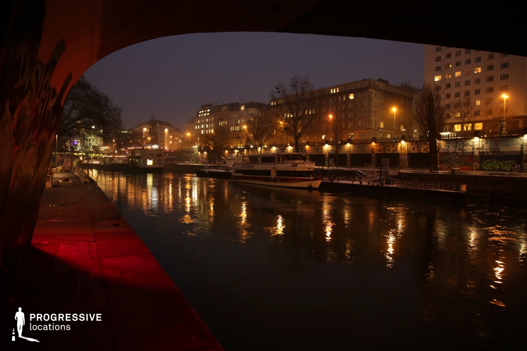 This moody riverside scene is framed by the underside of a bridge, adding a sense of depth and enclosure. The softly illuminated water reflects the warm glow of streetlights and the lights from surrounding historic buildings, creating a cinematic interplay of light and shadow. Graffiti-covered walls add a gritty, urban contrast to the otherwise elegant architecture. A docked boat enhances the sense of stillness, while distant city lights suggest activity beyond the immediate scene. The composition is ideal for noir-inspired films, crime thrillers, or contemplative dramas, offering a visually rich setting that blends urban grit with melancholic beauty.
