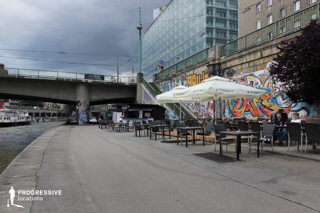 This riverside café terrace is set along a concrete embankment, juxtaposing industrial and modern aesthetics. Overhead, a sturdy bridge with tram cables spans the river, adding a raw, urban dynamic. The retaining wall, covered in vibrant graffiti, contrasts sharply with the sleek glass façade of a towering modern office building above. The café features simple outdoor seating with large branded umbrellas, creating a casual yet gritty atmosphere. The layered architectural elements, from the graffiti-covered steps to the contemporary high-rise, make this an ideal filming location for urban dramas, crime thrillers, or street-culture-inspired productions, offering an authentic metropolitan backdrop.