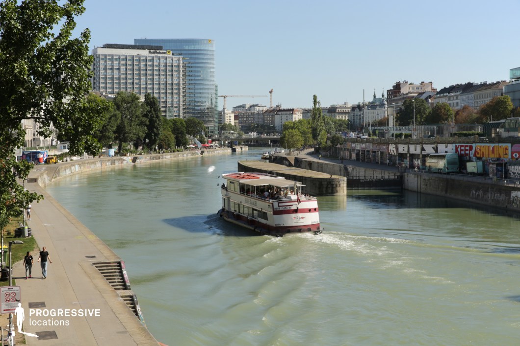 This riverside scene showcases a dynamic urban environment where modern architecture meets historical cityscapes. A sleek, glass-covered high-rise dominates the background, contrasting with classic European-style buildings lining the riverbank. A pedestrian walkway follows the gentle curve of the water, bordered by graffiti-covered walls that add a contemporary, artistic element. A boat navigates the waterway, emphasizing the city’s connection to the river. The mix of urban textures, from smooth concrete to greenery and reflective glass, creates a visually engaging setting. This location is ideal for films focusing on urban life, crime dramas, or establishing shots for metropolitan narratives, offering a balance between modern sophistication and raw street culture.
