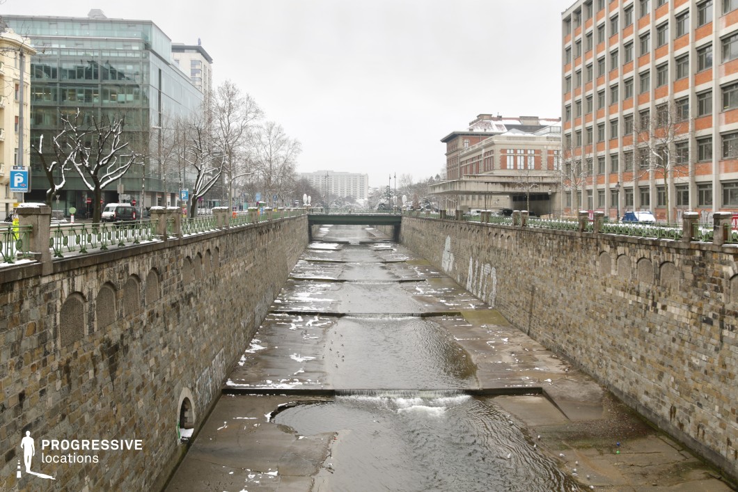 This structured urban canal is flanked by tall stone walls, creating a linear, enclosed setting with a strong sense of perspective. The waterbed is partially structured with concrete barriers, adding an industrial feel to the otherwise historical masonry. Above, a mix of modern glass buildings and traditional brick facades line the street, offering a contrast of architectural styles. Decorative railings and evenly spaced stone pillars top the embankments, reinforcing the symmetrical depth of the scene. The combination of rigid geometry and muted tones makes this location ideal for crime dramas, neo-noir thrillers, or dystopian settings, offering a stark and atmospheric urban backdrop.