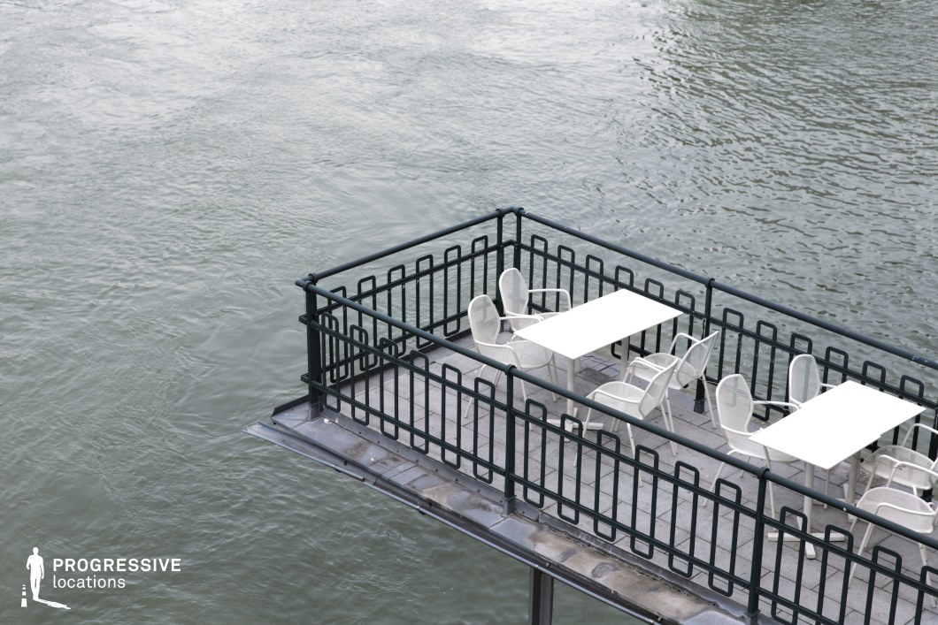 This suspended riverside terrace features a minimalist design with simple white tables and chairs enclosed by a dark metal railing, extending over the water for an immersive, tranquil setting. The calm, textured surface of the river dominates the background, creating a sense of isolation and intimacy. The contrast between the stark geometric lines of the terrace and the organic movement of the water enhances the visual appeal. This location is ideal for introspective or romantic scenes, stylish commercial shoots, or films exploring solitude, reflection, and urban serenity.