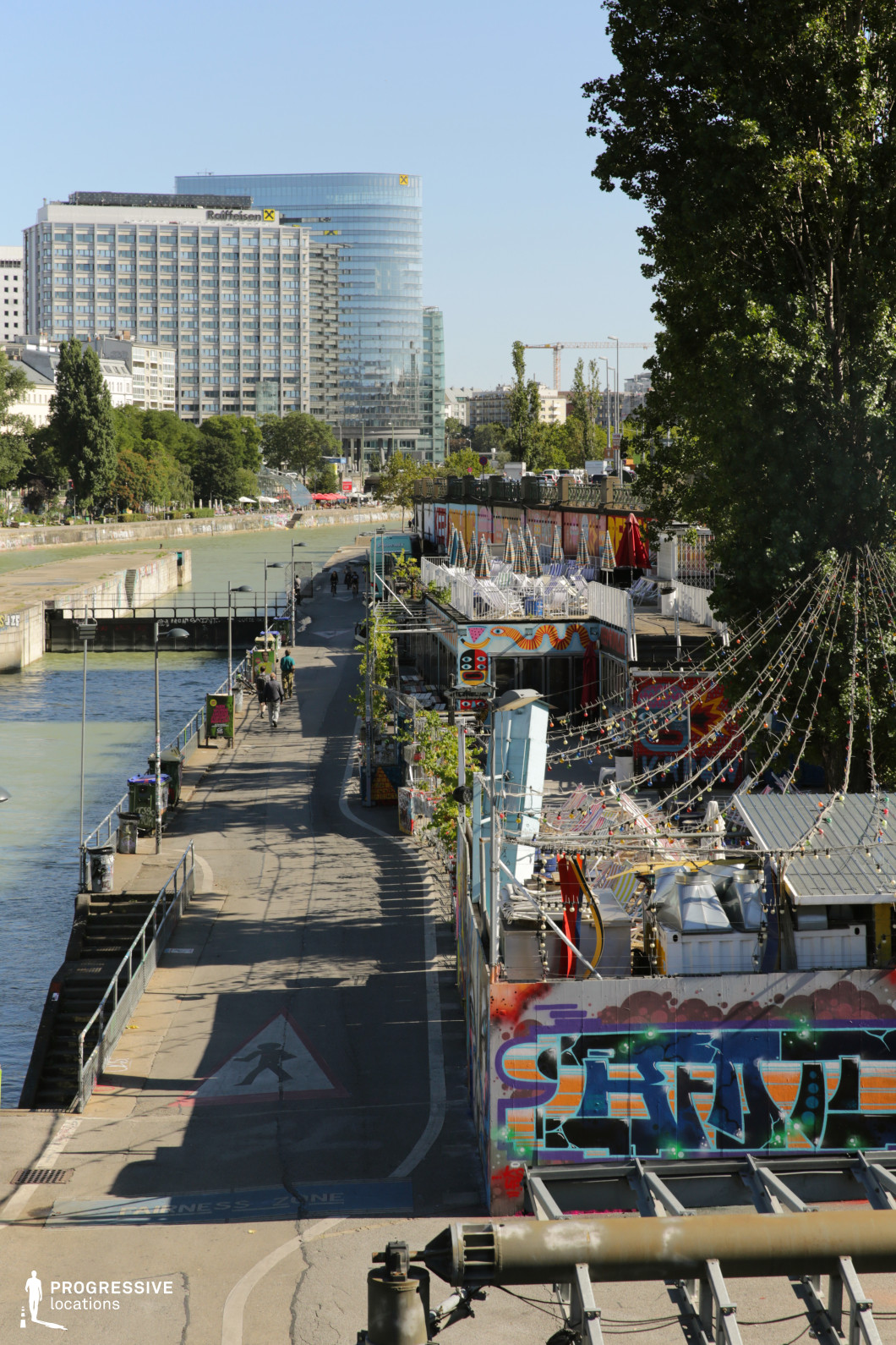 This dynamic urban riverside scene features a canal walkway lined with colorful graffiti-covered walls, casual outdoor bars, and string lights, creating a vibrant contrast against the sleek modern high-rises in the background. The paved promenade runs parallel to the water, with metal railings and lampposts adding structure to the scene. The eclectic mix of street art and contemporary architecture gives the location an edgy, metropolitan feel, perfect for filming urban dramas, music videos, or counterculture narratives. The depth of the perspective, with the canal stretching into the distance, also makes it ideal for visually striking tracking shots and atmospheric city montages.