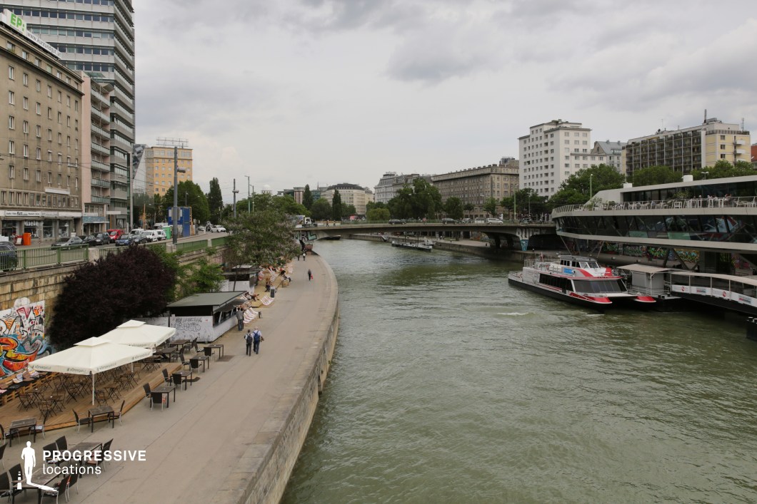 This lively urban riverside setting combines modern infrastructure with relaxed social spaces. A gently curving concrete embankment hosts outdoor cafés, deck seating, and graffiti-adorned walls, creating an informal yet dynamic waterfront atmosphere. The river, lined with contemporary floating structures and boats, is spanned by multiple bridges connecting different parts of the city. Surrounding buildings showcase a mix of older European architecture and modern high-rises, emphasizing the evolving cityscape. This location is ideal for filming contemporary dramas, crime thrillers, or travel and lifestyle segments, offering a visually rich backdrop that blends leisure, movement, and urban energy.