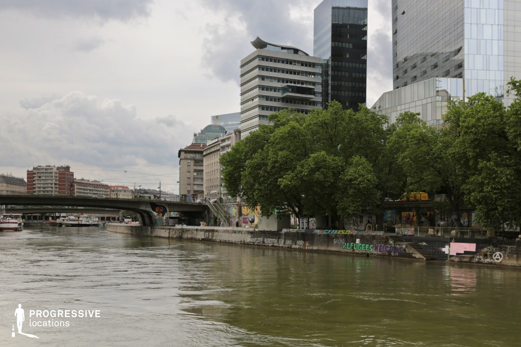 This dynamic riverside location blends raw urban character with sleek modernity, featuring a mix of high-rise glass buildings, mid-century architecture, and a graffiti-covered embankment. A low, weathered concrete bridge spans the water, adding an industrial touch to the scene. Lush trees along the riverbank contrast with the rigid structures, softening the city's edge. The presence of street art and makeshift elements suggests an alternative cultural space, making this setting ideal for gritty dramas, crime thrillers, or contemporary urban narratives. The layered architectural styles and reflective water surface enhance the cinematographic depth, perfect for scenes involving underground movements, introspection, or urban exploration.