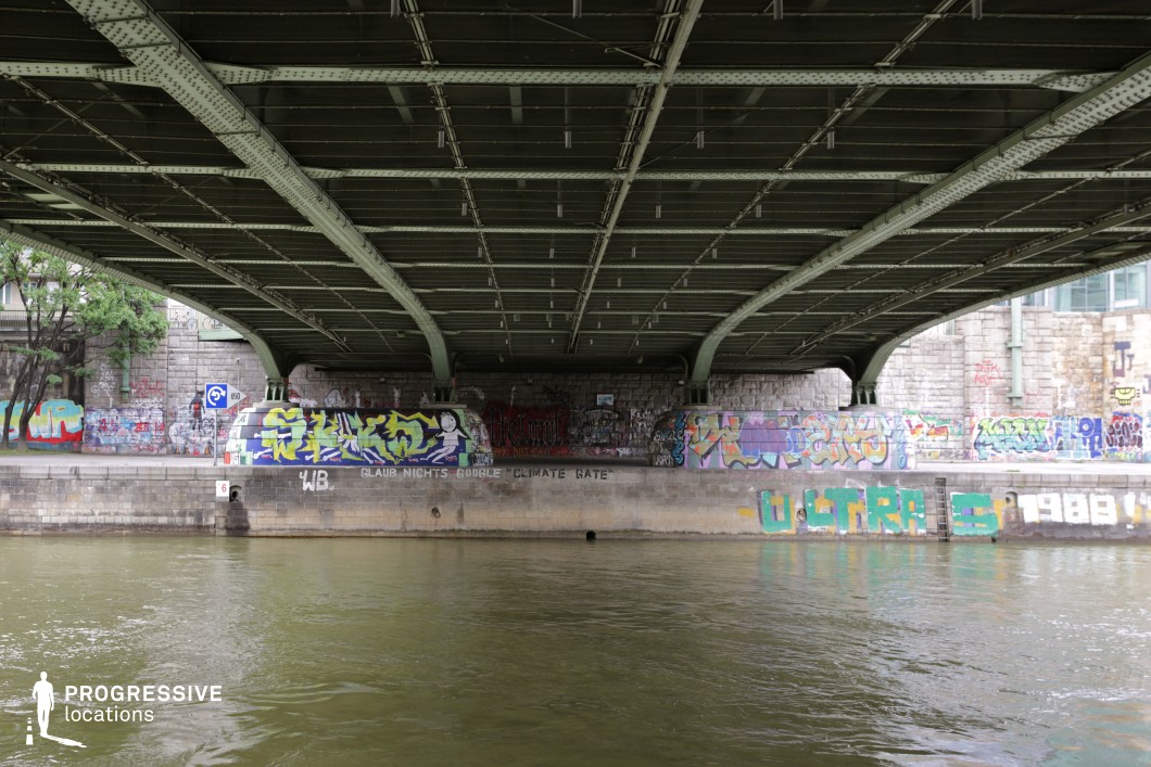 This urban riverside location features the underside of a steel bridge with industrial-style metal beams forming a rigid, structured ceiling. The concrete embankment beneath is covered in vibrant graffiti, creating a contrast between the cold, utilitarian architecture and the colorful, rebellious street art. The river flows calmly along the edge, adding a sense of movement and reflection to the scene. The stone walls and metal framework provide a gritty, urban aesthetic, making this setting ideal for crime dramas, action sequences, or underground subculture narratives. The layered elements of industrial infrastructure, street art, and water create a dynamic and visually rich backdrop for cinematic storytelling.