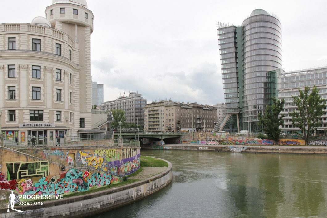 This dynamic riverside scene showcases a striking blend of historic and contemporary architecture. On one side, a grand neoclassical building with a rounded tower and ornate detailing stands as a reminder of the city's architectural heritage. Opposite, a sleek glass high-rise with curved lines and a futuristic design embodies modern urban development. The riverbanks are lined with graffiti-covered walls, adding an element of raw street culture that contrasts with the structured elegance of the buildings. The arched bridge and waterfront setting create depth and perspective, making this location ideal for filming urban dramas, crime thrillers, or narratives that explore themes of gentrification, subcultures, and city life.