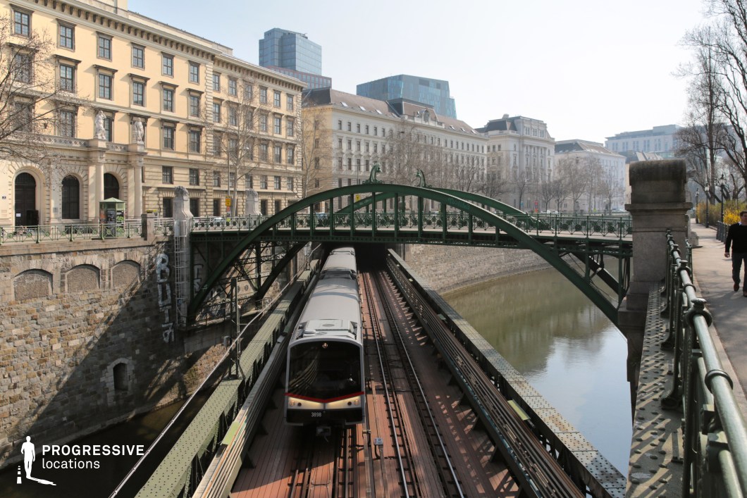 This striking urban scene features a green iron bridge arching over a railway line, set against a backdrop of classical and modern architecture. The railway runs through a stone-lined canal, creating a dynamic contrast between industrial functionality and historic elegance. The bridge's intricate metalwork and pedestrian walkway add depth and movement to the composition, while the gently curving waterway enhances the visual balance. The mix of old-world facades and contemporary high-rises in the background creates a layered cityscape. This location is ideal for filming urban dramas, crime thrillers, or high-paced action sequences, offering a rich setting for moments of transit, pursuit, or introspective city walks.