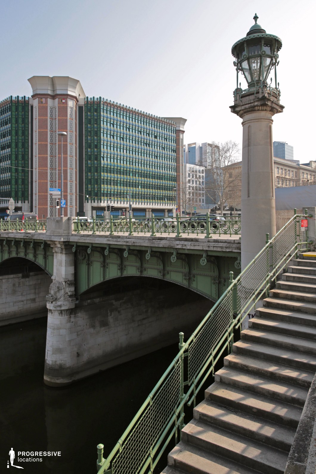 This location features an ornate green metal bridge with intricate detailing, standing over a calm waterway. A grand stone staircase with matching green railings leads down to the lower embankment, creating strong geometric lines and depth. A tall, decorative lamppost with a historic design adds an elegant focal point. In contrast, the background showcases a mix of architectural styles, including a bold modernist building with patterned facades and glass windows, alongside traditional structures. The interplay of classic and contemporary elements makes this setting ideal for sophisticated urban dramas, espionage thrillers, or architectural documentary projects, offering rich visual contrast and layered storytelling potential.