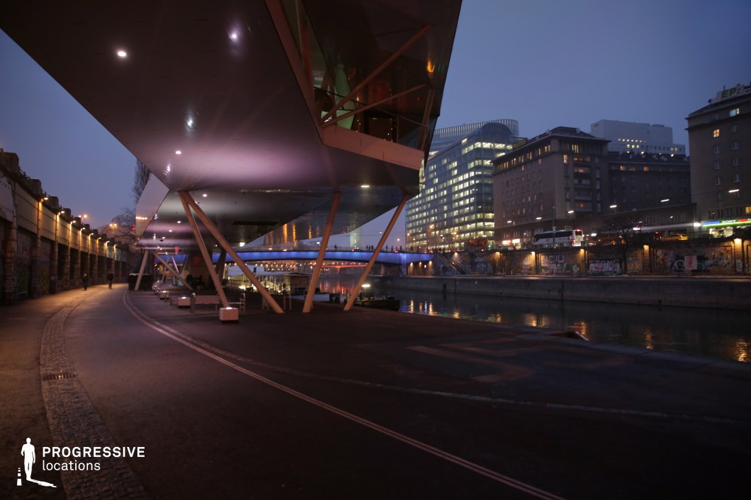 This modern riverside location features a striking architectural structure with an angular overhang, supported by slender steel beams, casting a dramatic presence over the wide pedestrian walkway. Soft purple and white lighting under the canopy enhances the futuristic ambiance, contrasting with the illuminated city skyline in the background. The river reflects the glow of the bridge and surrounding buildings, adding depth and dynamism to the scene. Graffiti-covered walls along the path contribute an urban, gritty edge, making this an ideal setting for neo-noir thrillers, cyberpunk-inspired narratives, or contemporary dramas. The mix of sleek design, industrial elements, and urban night lighting offers a visually compelling backdrop for high-stakes, atmospheric storytelling.