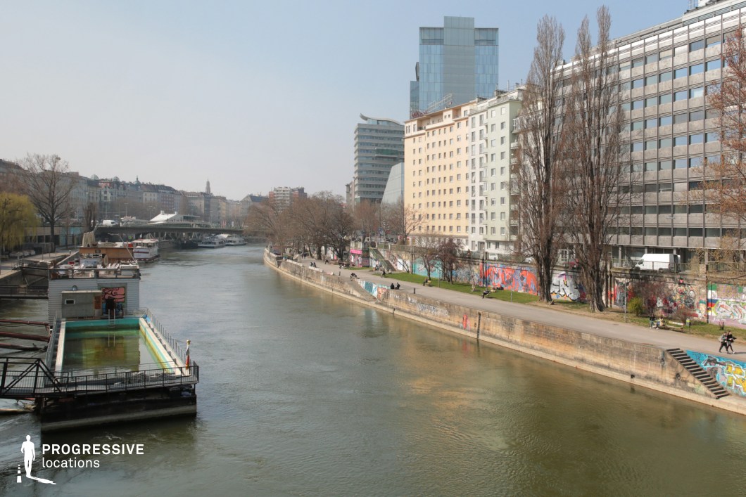 This image captures a dynamic urban riverside scene where a mix of modern glass skyscrapers and mid-century residential buildings line the waterfront. The river flows between graffiti-covered embankments, creating a striking contrast between the artistic, raw aesthetic of the walls and the structured, contemporary architecture. A floating structure with an unused pool in the foreground adds an industrial and slightly neglected feel, reinforcing the setting’s urban edge. The perspective leads the eye along the river toward a bridge and additional city structures, creating depth. This versatile location is ideal for filming urban dramas, crime thrillers, or scenes involving solitude, movement, or transformation within a metropolitan environment.