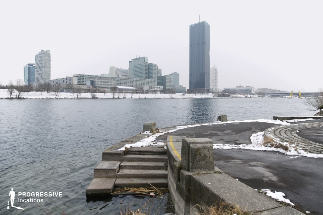 This riverside location combines raw, industrial elements with a striking modern cityscape. In the foreground, a worn concrete platform with descending steps leads to the water’s edge, adding a rugged, utilitarian character. The dark pavement and geometric patterns of the walkway create a structured yet slightly abandoned feel. The background features a cluster of sleek high-rises, including a prominent dark glass skyscraper, giving the scene a distinctly corporate and futuristic look. The mix of natural water elements and urban infrastructure makes this site ideal for filming crime dramas, neo-noir thrillers, or dystopian sci-fi settings, offering a blend of isolation and contemporary urban tension.
