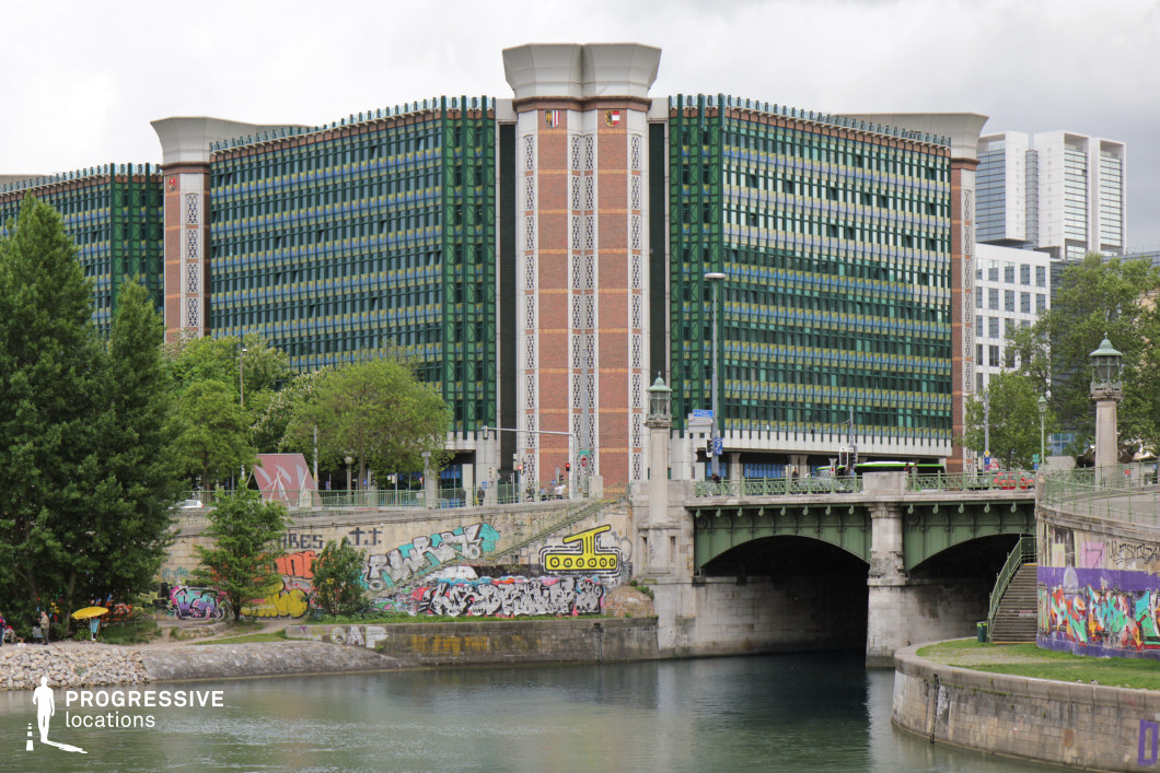 This urban waterfront setting presents a dynamic contrast between a striking modern glass-and-brick building, an ornate green metal bridge, and a graffiti-covered embankment. The structured, geometric facade of the contemporary architecture adds a formal, institutional quality, while the graffiti-adorned walls and stairways along the canal inject a raw, artistic energy. The waterway, framed by stone and metal railings, reflects the surrounding environment, enhancing the sense of depth and urban vibrancy. This location is well-suited for filming contemporary dramas, crime thrillers, or street-culture documentaries, offering a blend of corporate modernity, historic infrastructure, and underground artistic expression.